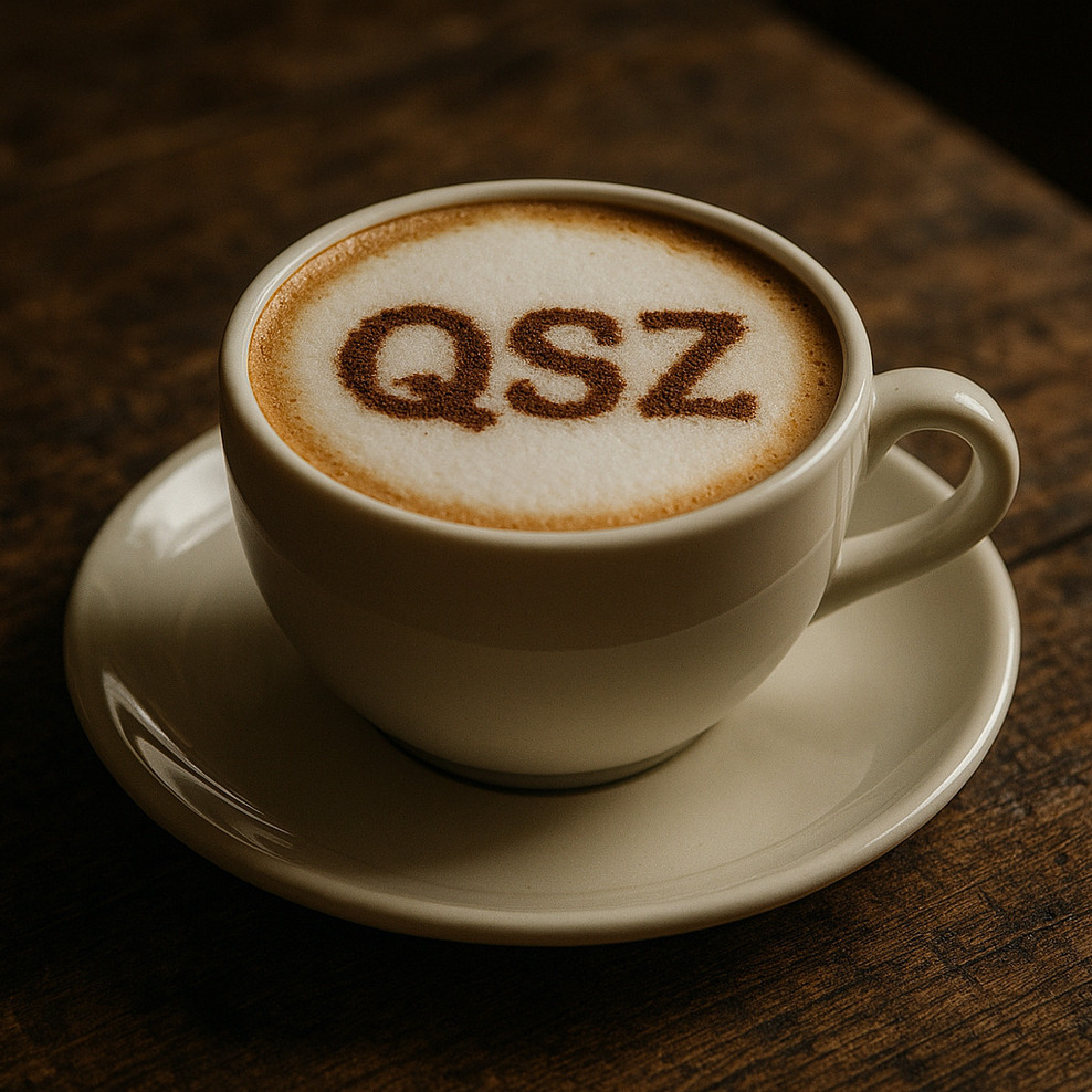 Eine Tasse Kaffee auf einem dunklen Holztisch, in der Tasse Milchschaum, auf den mit Kakao die Buchstaben QSZ gestreut sind.